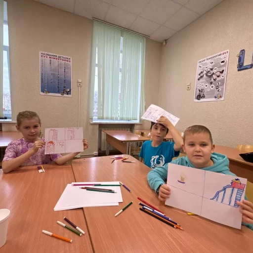 Трое детей на занятии в Лиге Роботов (Екатеринбург) показывают эскизы будущих роботов; на столе цветные карандаши, на стене постеры EV3